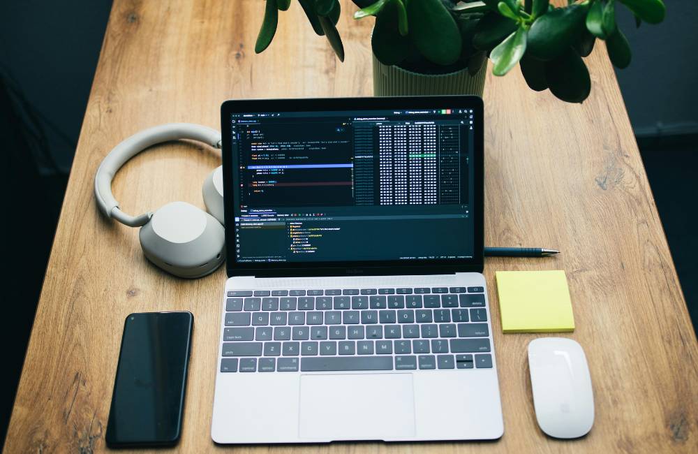 明るい木目のデスクに、開いたノートパソコン、スマホ、白いワイヤレスマウス、黄色い付箋、黒いペンが整然と並べられている上からのショット。シンプルで整理されたワークスペースの様子。