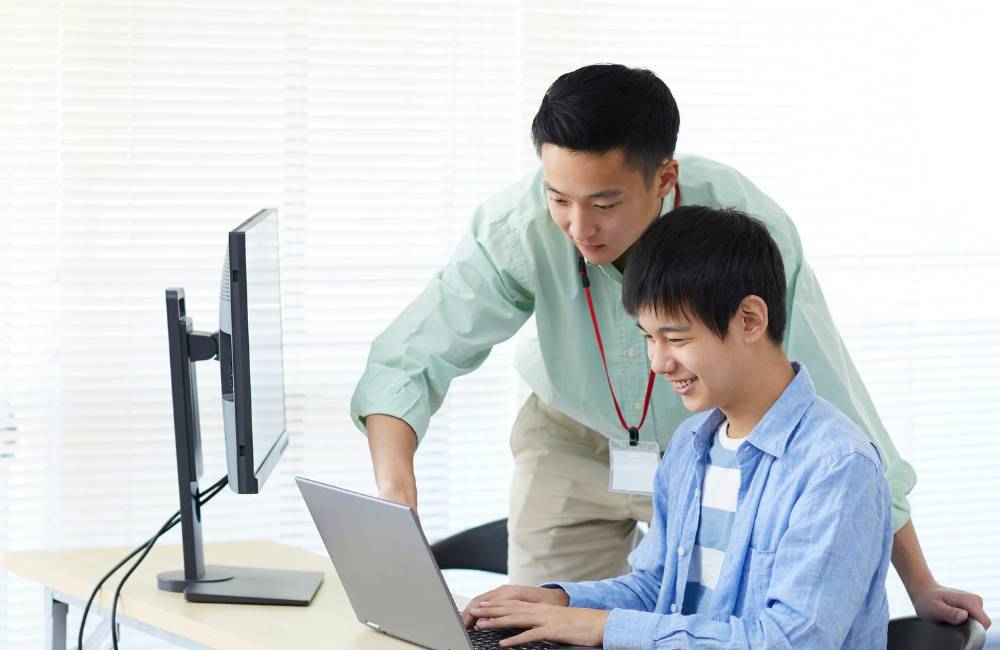 男性が隣に座る少年にパソコンの画面を指差しながら、笑顔で使い方を教えているシーン。中古パソコン選びの不安が解消され、前向きな気持ちで使い始める様子を表現しています