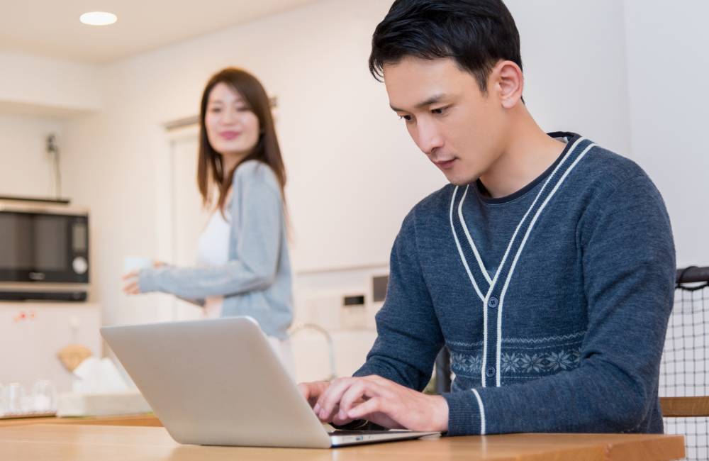 リビングのテーブルで、家族の気配を感じながら笑顔でノートパソコンを操作する男性の様子