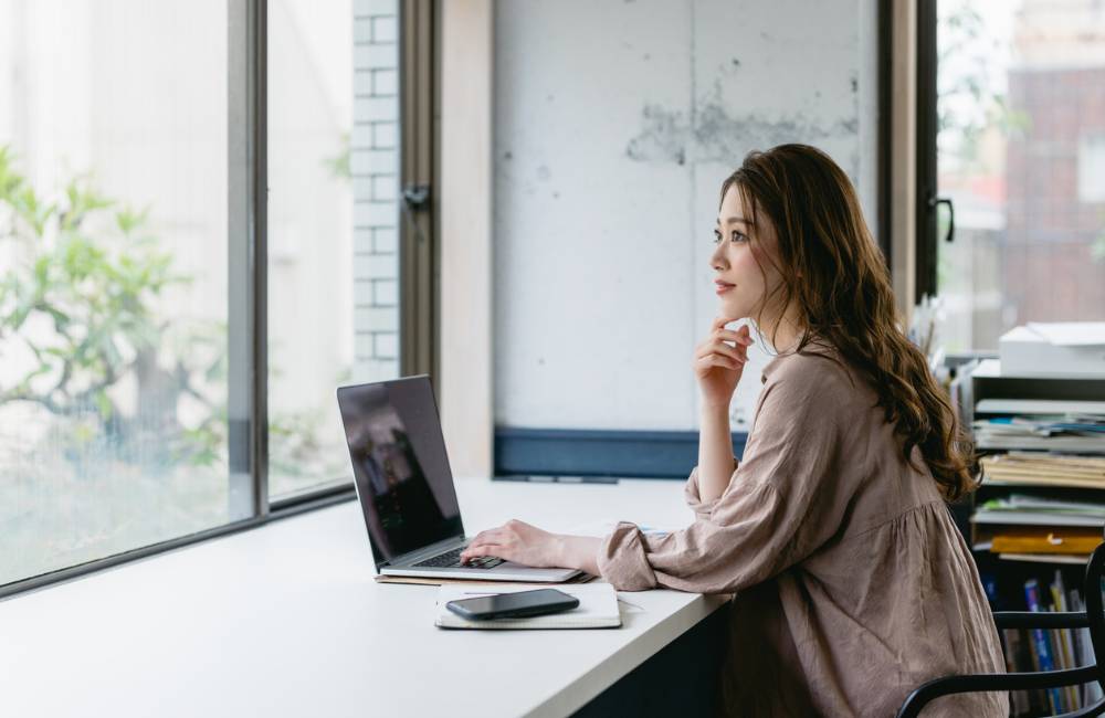 明るい窓際のデスクで、ノートパソコンを前にして「どの1台にしようか」と指をあごに当てて考え事をする女性の横顔