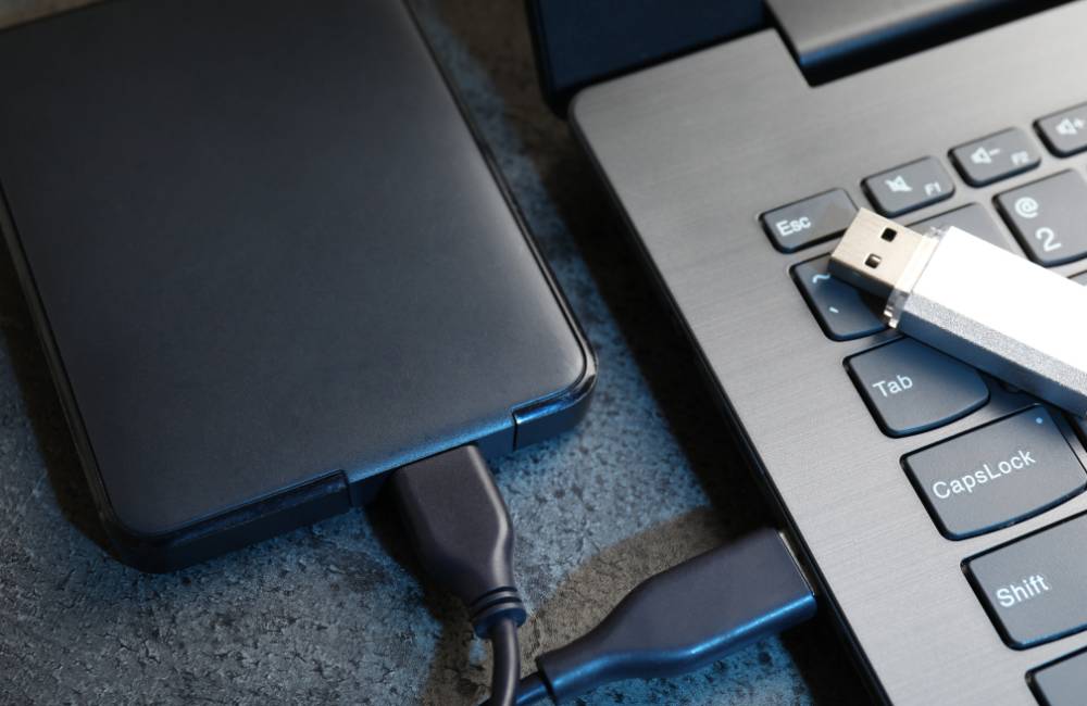 ノートパソコンの側面に外付けハードディスクを接続し、キーボードの上に置かれたUSBメモリを確認している接写写真