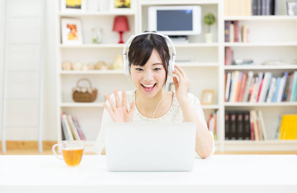 白いヘッドホンをつけ、ノートパソコンの前で笑顔で手を振る女性。背景には本棚があり、自宅で楽しくオンライン交流をしている様子。