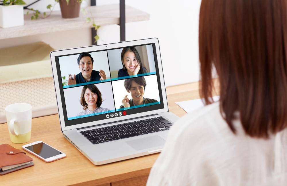 ノートパソコンの画面越しに、4人の男女が笑顔でビデオ会議を行っている様子。明るく信頼感のあるコミュニケーションの風景。