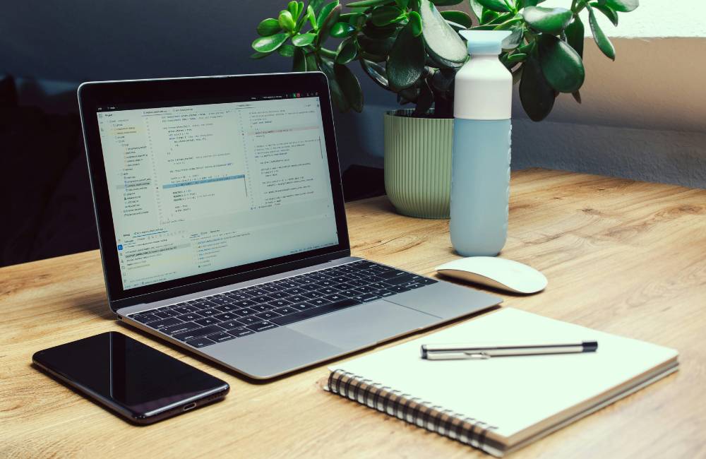 木目調のデスクに、プログラムコードが表示されたノートパソコン、スマホ、ノート、筆記用具、水筒が整然と並んでいる仕事場。