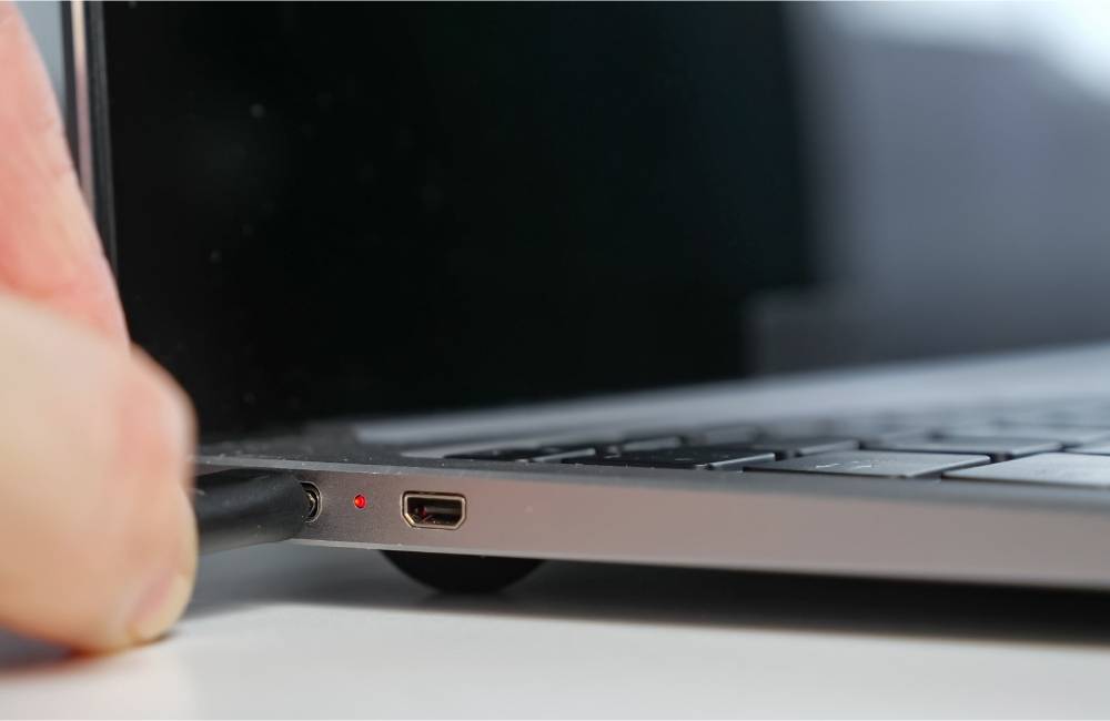 ノートパソコンの横にある小さな充電ポートに、黒い充電ケーブルの先端をゆっくりと差し込もうとしている様子。