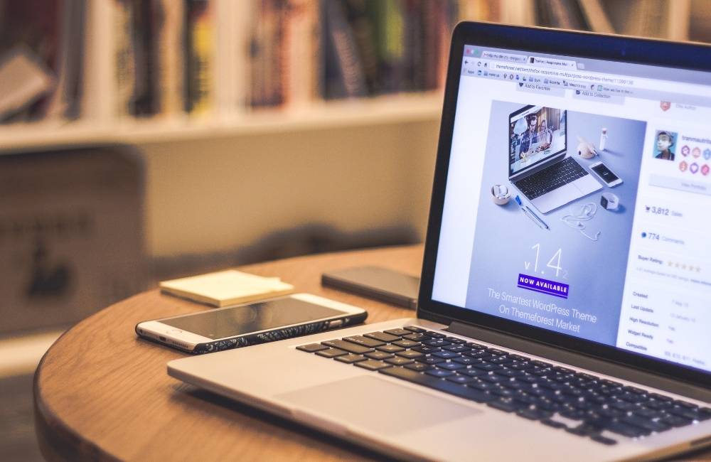 木目のデスクに置かれたノートパソコンの隣に、画面を上にしたスマートフォンが置かれ、落ち着いて作業している風景。