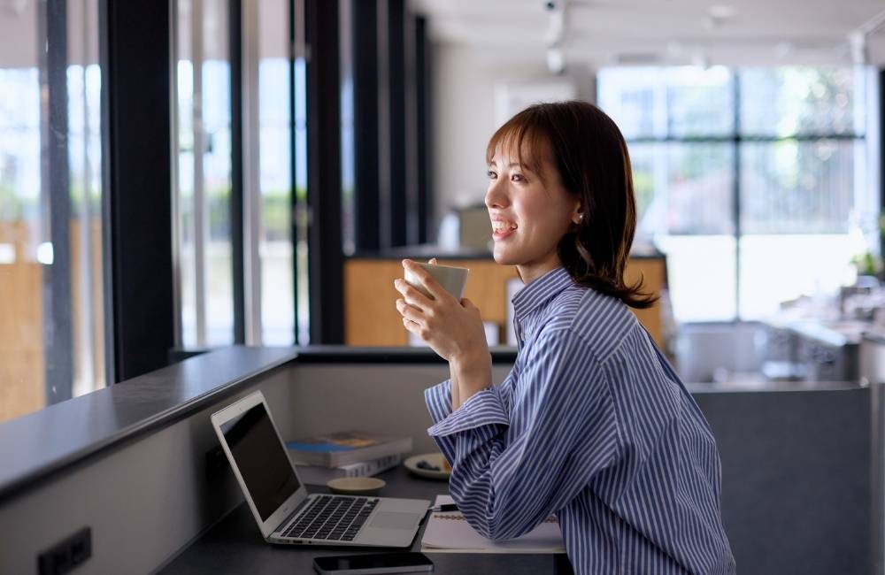 窓際の明るい席で、温かい飲み物を片手に笑顔でノートパソコンを眺めている、リラックスした女性の横顔。