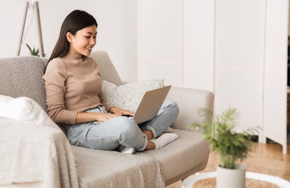 お部屋のソファでクッションを背中に、リラックスした姿勢で膝の上にパソコンを置いて楽しそうに操作している女性。