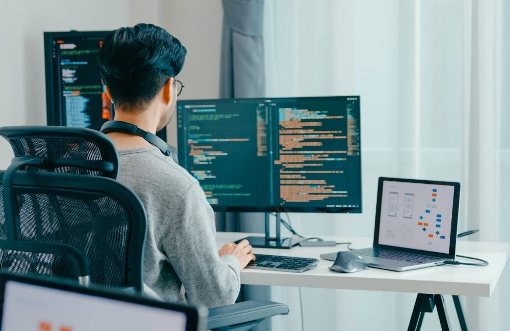 デスクに並んだ2枚のモニターに、英語のプログラムや図面をたくさん表示して作業している後ろ姿