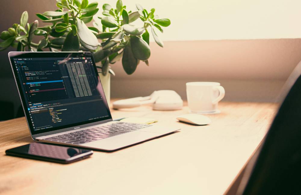 木目のデスクで、プログラミング用の複雑なコードが表示されているノートパソコン。