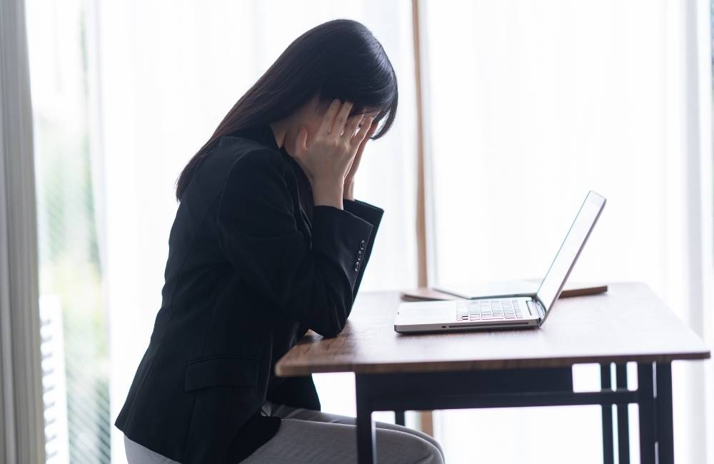 パソコンがうまく動かず、机に両肘をついて頭を抱えながら悩んでいる女性の様子。