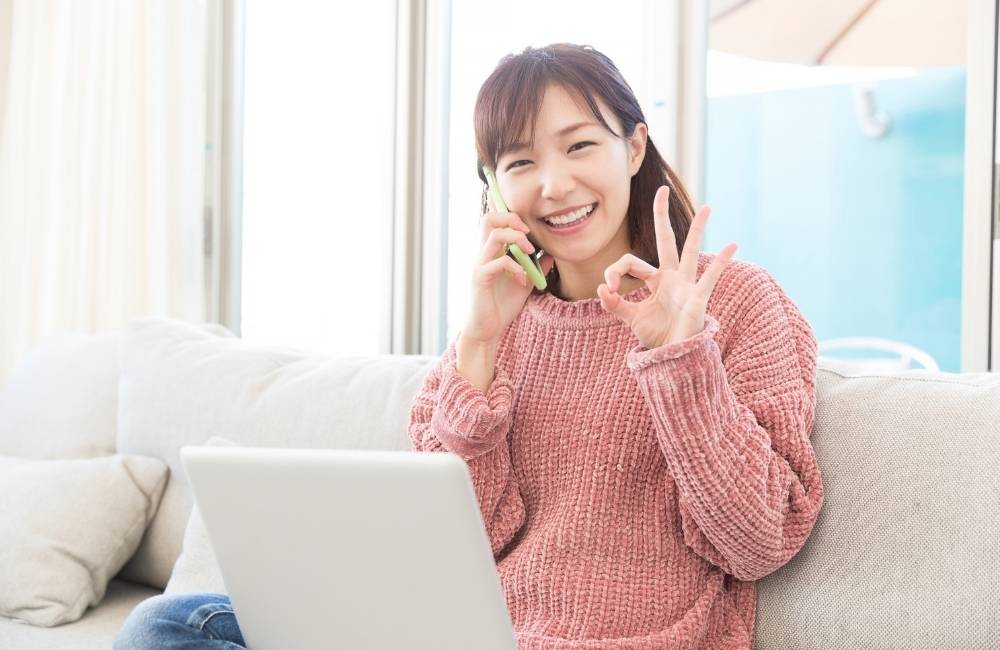 リビングのソファでノートパソコンを使いながら、笑顔で電話をして「大丈夫」とOKサインを出している女性。