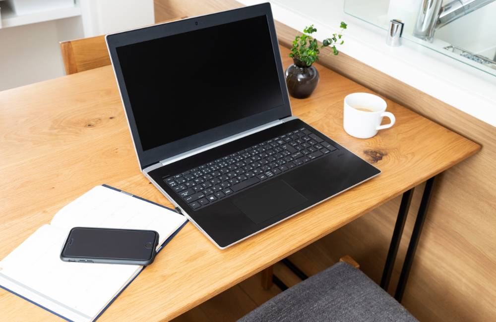 明るい木目のデスクに、開いたノート、スマートフォン、黒いノートパソコン、そして温かい飲み物が入ったマグカップが整然と並んでいる様子。購入前に頭の中をスッキリ整理するイメージにぴったりです。