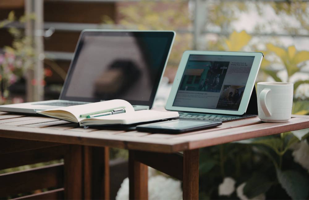 屋外のテーブルで、ノートパソコンとタブレット、コーヒーを置いて作業している穏やかな風景。