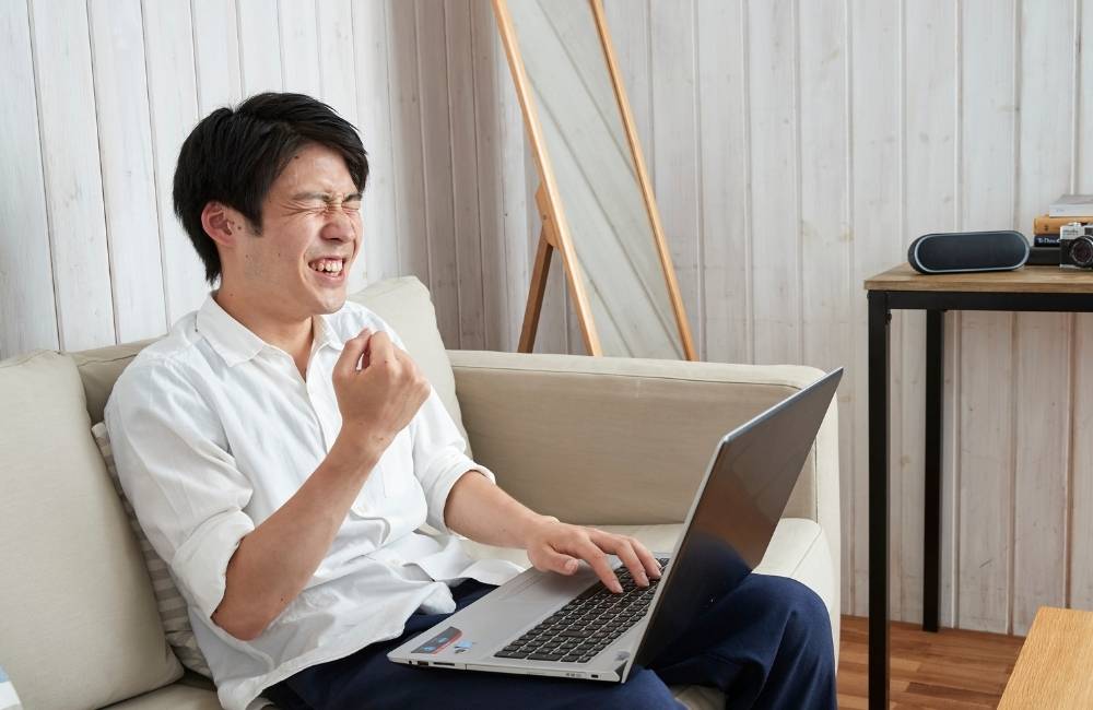 ソファでノートパソコンを使いながら、インターネットが無事に繋がって嬉しそうに拳を握り、ガッツポーズをする男性。