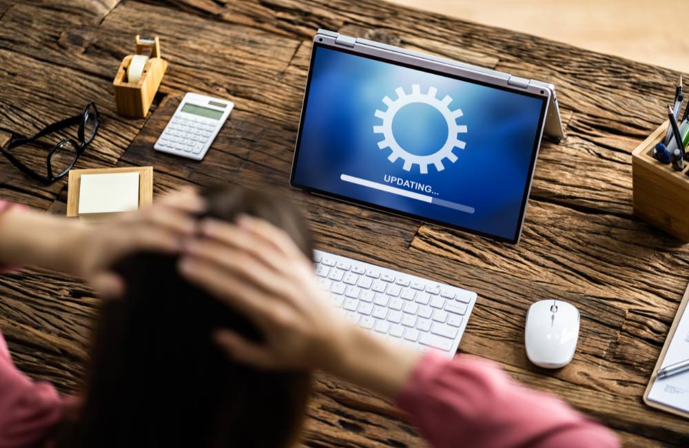 古い木製の机に置かれたノートPCが「UPDATING...」と表示されている。その後ろで女性が不安そうに頭を抱え、アップデートの待ち時間と心配な気持ちを表現している。