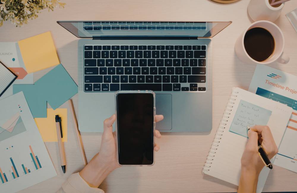 ノートパソコン、スマホ、コーヒー、メモ帳が整然と並んだデスクで、ペンを使ってタスクを書き留めている整理された作業環境