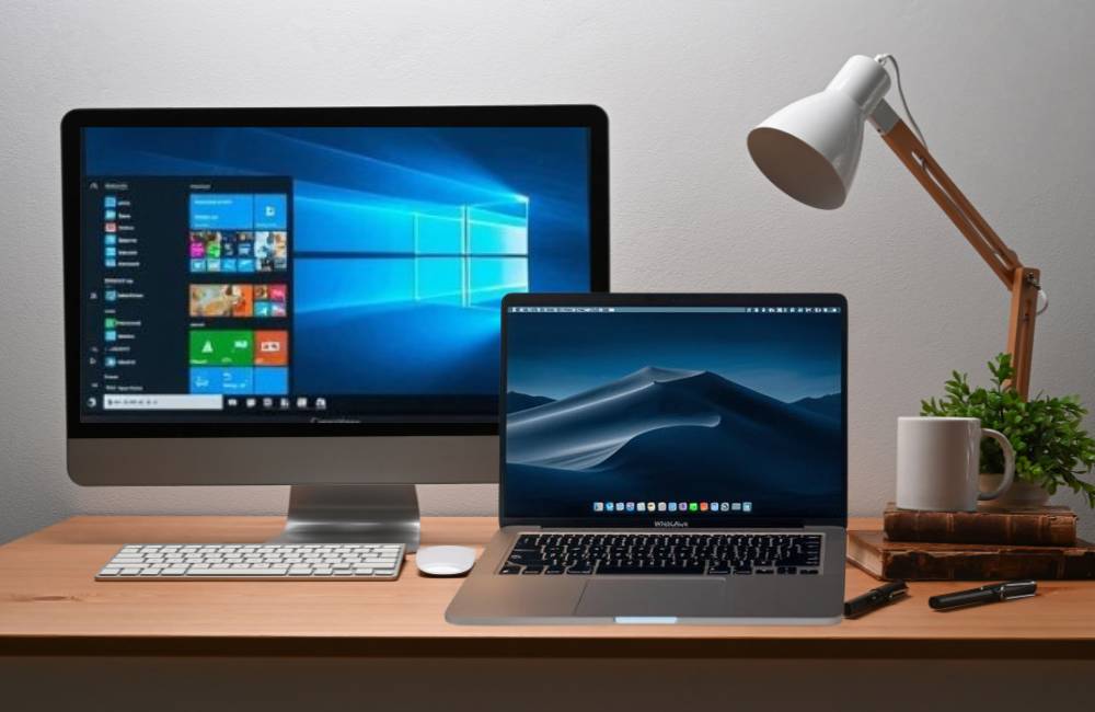 デスクの上にWindowsのOS画面が表示された一体型PCとMacBookが並べて置かれている様子