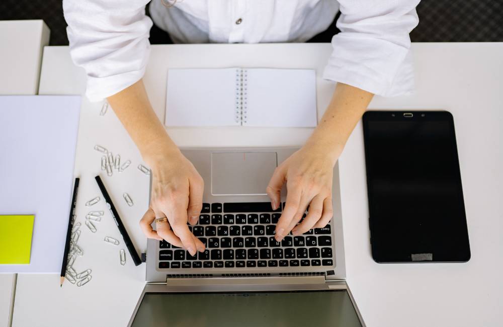 白いデスクの上でノートパソコンのキーボードを両手でタイピングしている俯瞰写真