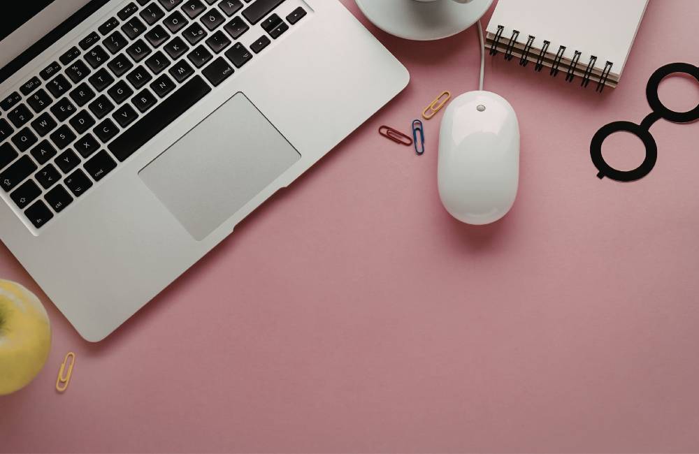 ピンク色のデスクに配置されたノートパソコンの一部と白いマウス、丸メガネの小物