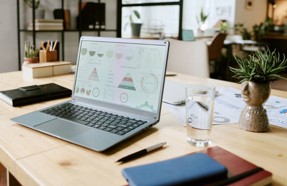 木製のデスクに置かれた、シルバーのノートパソコンの画面。円グラフや棒グラフ、表など、カラフルなビジネスデータが表示されている。その隣には水の入ったグラスと緑の植物があり、HPのパソコンがビジネスの現場で活躍している様子をイメージさせる。