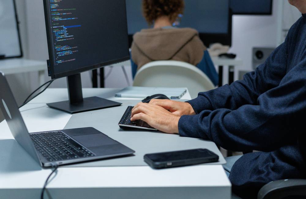 暗い部屋で、ノートパソコンと大きなモニターの前に座るユーザー。モニターには、青と緑の文字でプログラミングコードやセキュリティログのようなデータが表示されており、HPのビジネスモデルが持つ強力なセキュリティ機能を象徴している。