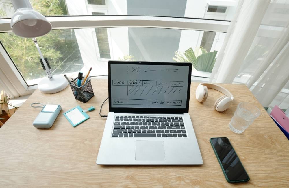 木目調のデスクに置かれたノートパソコンと、設計図のようなワイヤーフレームが表示された画面