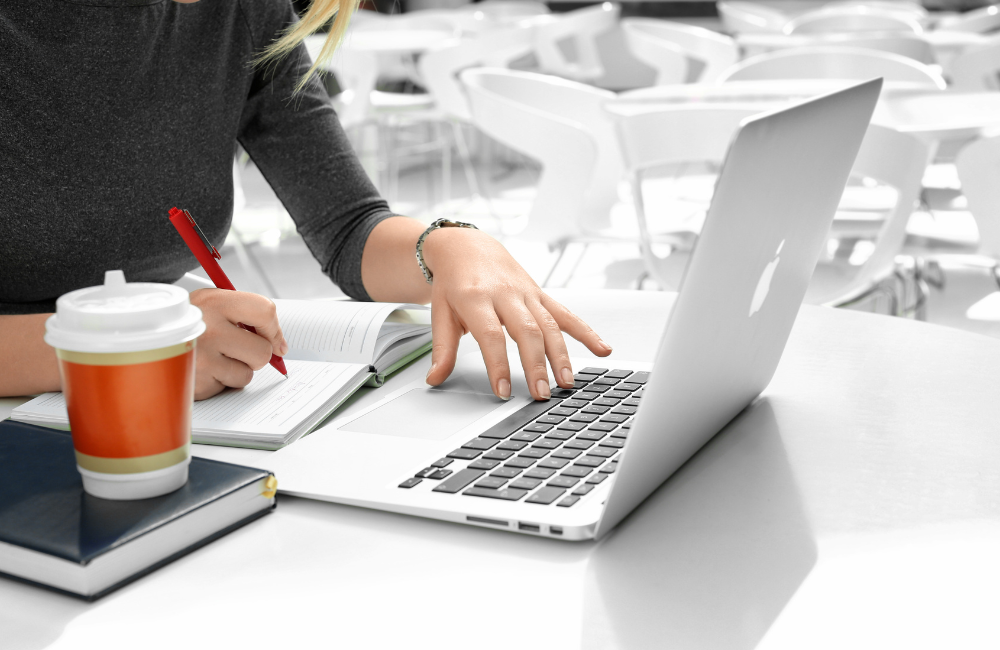 木目のデスク上で、開いたノートブックにペンを走らせる手元と、その横に置かれたMacBookのクローズアップ。