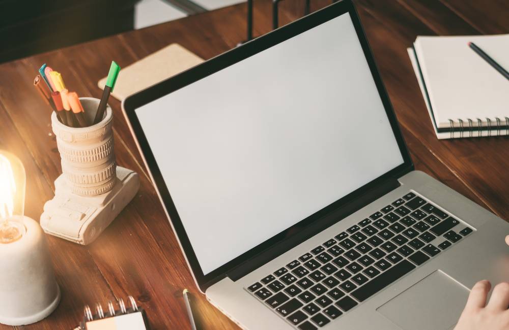 木製のデスクに、ノートパソコン、ノート、ペン、そして点灯した電球が置かれている。