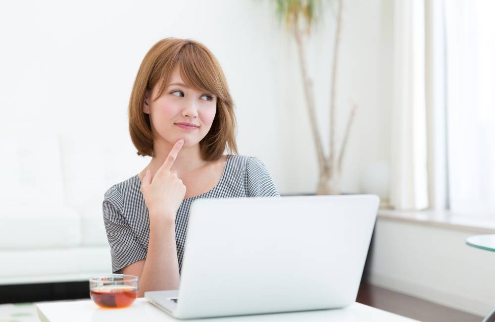 ノートパソコンの前に座り、人差し指を顎にあてて思案する女性。