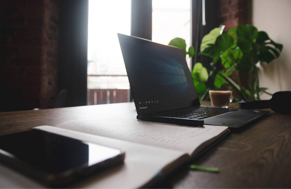 日当たりの良い窓際のテーブルの上で、ノートやスマートフォン、コーヒーと一緒に置かれているビジネス用ノートパソコン