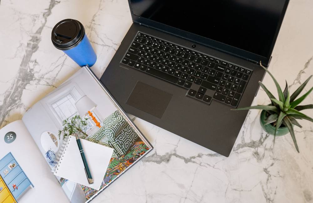 大理石風のテーブルの上にノートパソコン、雑誌、メモ帳、コーヒーが置かれた、洗練された作業環境の俯瞰写真