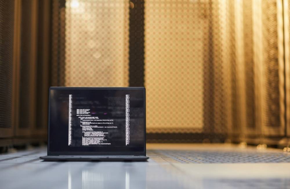 サーバーが並ぶ施設内で、画面にシステムコードが表示されたノートパソコンが置かれている様子。