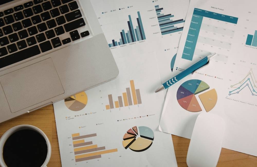 資料やグラフ、コーヒーカップが並ぶデスクで、ノートパソコンを開いてデータを確認している俯瞰写真。