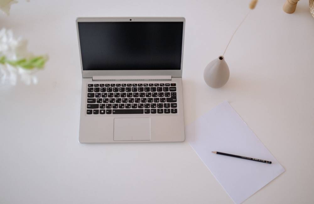 白いデスクに置かれたノートパソコンと、一輪挿しの花、鉛筆、白い紙が整然と並ぶシンプルな風景。