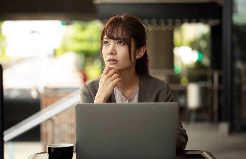 屋外のカフェでノートパソコンを前に、不安そうな表情で顎に手を当てて悩み込んでいる女性の姿