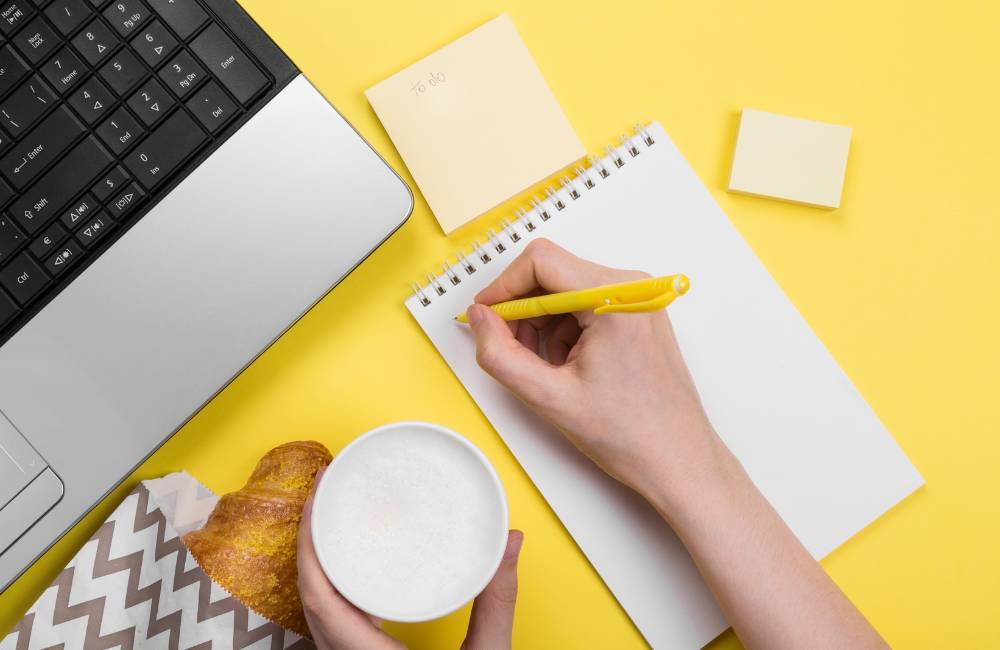 黄色の背景に、ノートパソコン、コーヒー、クロワッサン、そしてチェックリストを作成するためにペンでノートに書き込みをしている手元の俯瞰写真