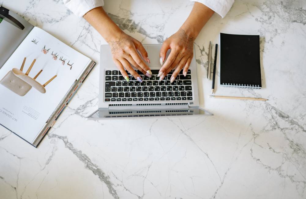 大理石のデスクでノートパソコンを操作しながら、雑誌やノートを広げて作業の準備を整える様子