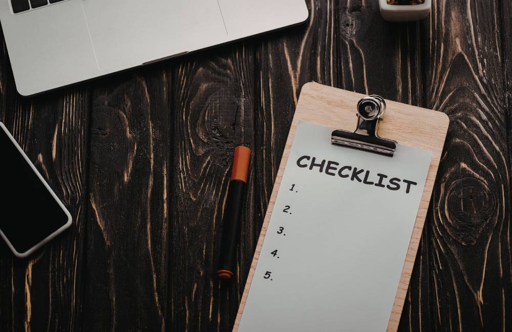 木目のデスクに置かれた、5つの項目が並ぶ空のチェックリストとノートパソコン